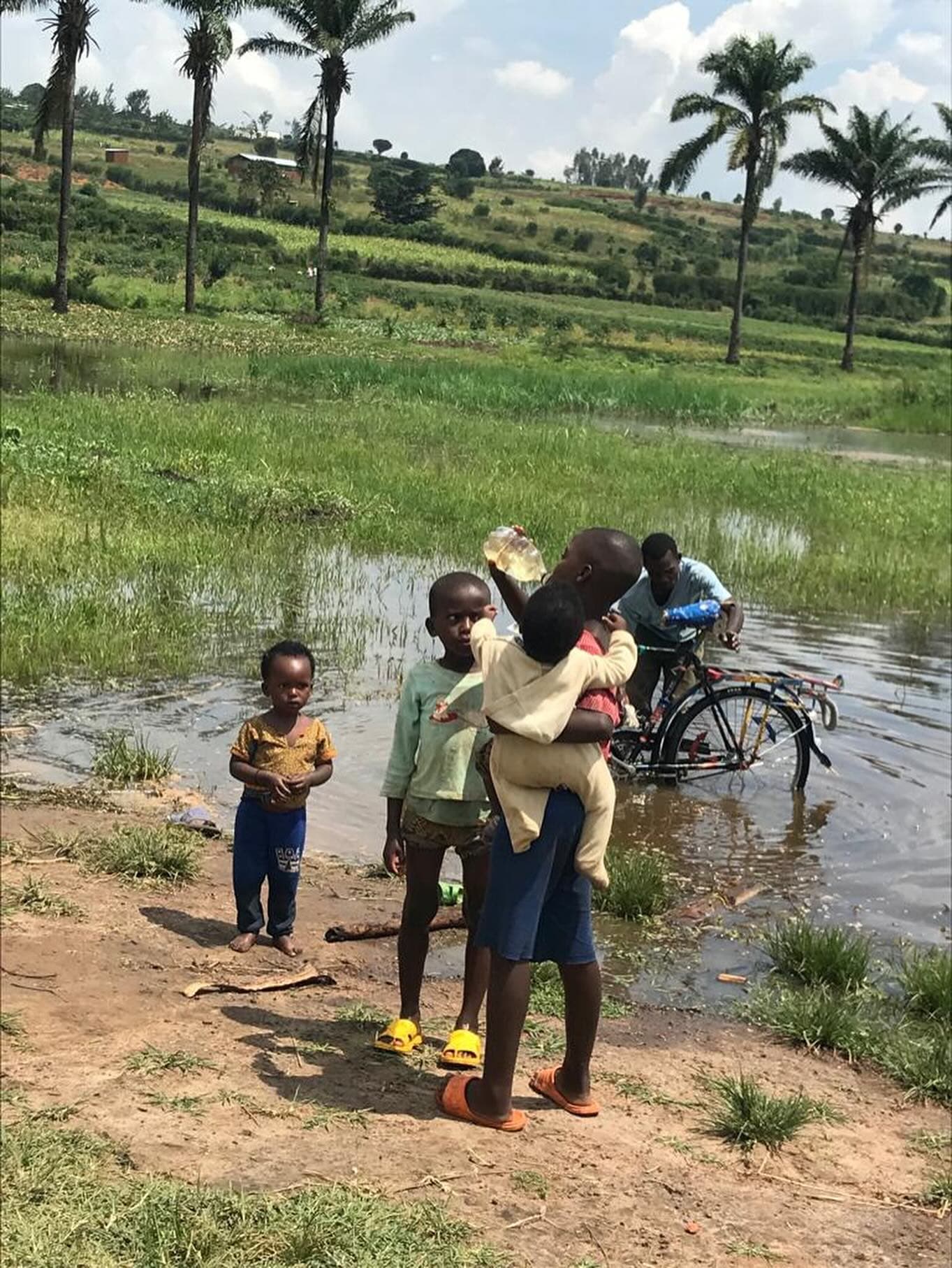 Children collecting water from Lake Mirayi while a man washes a bicycle in the water behind them.